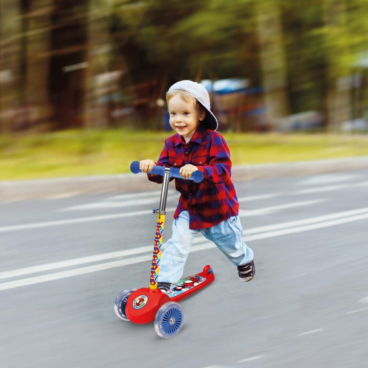 Little boy in cap is happily riding scooter at summer park backg